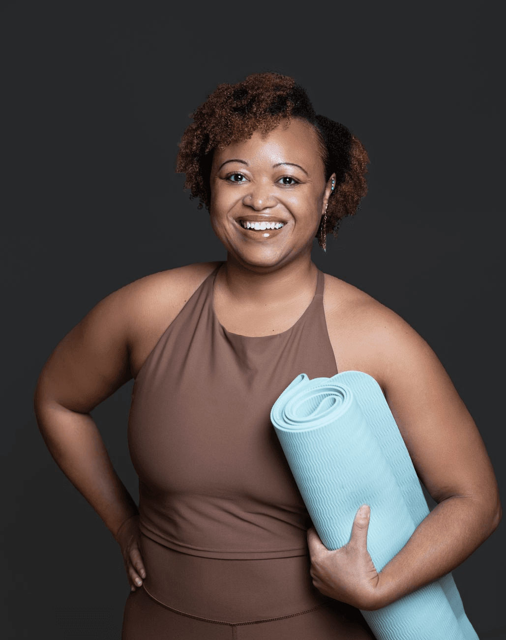 Woods Adisa, wellbeing facilitator and yoga instructor, standing with a rolled yoga mat, smiling.